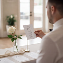 A man is seated at a table with a white rose in a vase, holding a paper with text on it.