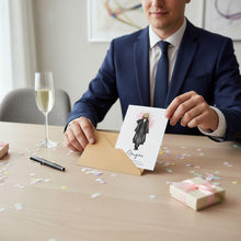 A man in a suit is sitting at a table, holding a card with a drawing of a person on it. There is a glass of champagne and confetti on the table, suggesting a celebratory occasion.