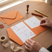 A person is arranging an orange wedding invitation on a wooden table, surrounded by stationery items such as envelopes, a wax seal, and a pen.