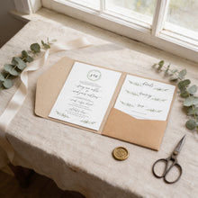 A wedding invitation card with a wreath and a small gold wax seal is placed on a table, accompanied by a pair of scissors and some greenery.