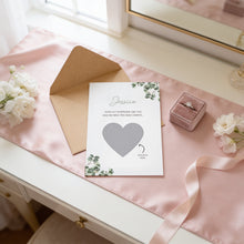 A wedding invitation card with a heart-shaped cutout, a pink velvet box containing a ring, and some white flowers on a pink tablecloth.
