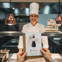 A person holding a card with an illustration of a chef in front of a chef in a kitchen, with a cake visible in the background.