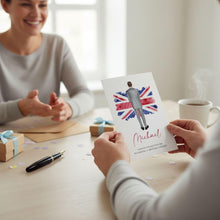A person is holding a card with a picture of a man wearing a suit and tie, with the text "Michael" written on it. The card is being held up in front of another person who is smiling and looking at the card. There are also some other items on the table, such as a pen, a cup, and a gift box.