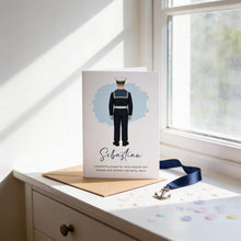 A card with a picture of a person in a military uniform, likely a sailor, and the name "Sebastian" printed on it. The card is placed on a white surface, with a blue ribbon and scattered confetti around it.