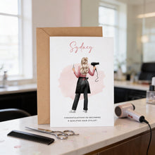 A greeting card with an illustration of a woman holding a hairdryer, celebrating her becoming a qualified hair stylist. The card is placed on a counter with a hairdryer, scissors, and a comb.