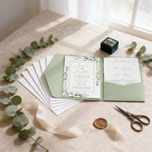 A wedding invitation with a green and white color scheme, featuring a floral design, placed on a table with a ribbon, a ring box, and some greenery.