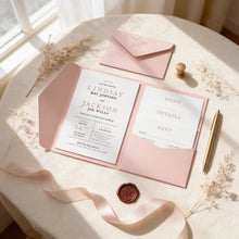 A pink wedding invitation card with the names Lindsay and Jackson, along with details about their wedding, is placed on a table with a pink ribbon, a gold pen, and some dried flowers.