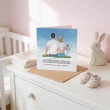 A greeting card with a couple sitting on a bench, with a baby in their arms, celebrating the birth of a baby named Andrews. The card is placed on a white dresser, with a pair of baby shoes and a white rabbit figurine nearby.