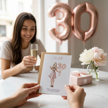 A woman is holding a birthday card with a design of a woman and the number 30, while another person is holding a champagne glass and a gift box. There are pink balloons and flowers in the background, and a pink gift box on the table.