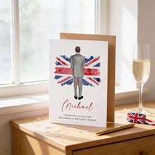 a card with a man standing in front of a Union Jack flag, and the card is placed on a wooden surface next to a glass of champagne and a pen.