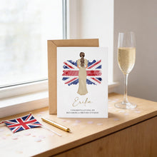 a card with a woman's portrait and a Union Jack flag, placed on a wooden surface next to a glass of champagne and a pen.