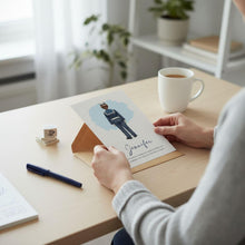 A person is sitting at a desk, holding a card with an illustration of a person and the name 'Jennifer' on it. There is also a cup of coffee and a pen on the desk.