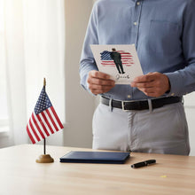 A person is standing at a desk, holding a card with an American flag design and a picture of a man on it. The desk also has a notebook, a pen, and a small American flag.