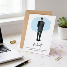 a greeting card with a man in a suit standing in front of a blue background, and the card is placed on a desk with a laptop, pen, and some confetti.