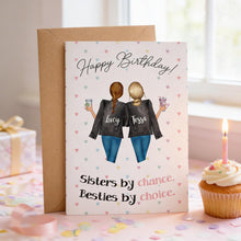 A birthday card with two women's backs facing each other, wearing matching black tops and blue jeans, with the text "Happy Birthday!" and "Sisters by chance, Besties by choice." The card is placed next to a cupcake with a lit candle, and confetti is scattered on the table.